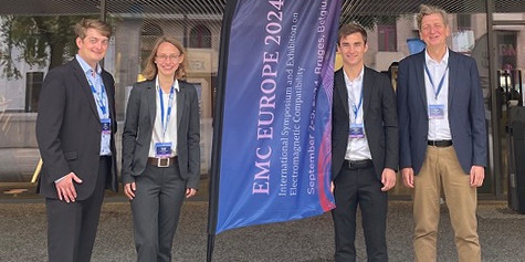 Drei wissenschaftliche Beschäftigte und Professor Frei sind auf dem Bild zu sehen. Sie stehen neben einer Fahne mit dem Schriftzug "EMC Europe 2024".